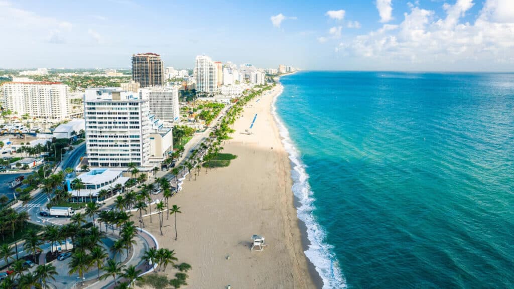 Aerial view of a sunny Miami beach with clear blue water, palm trees, and beachfront buildings.